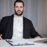 A serious bearded man in a black suit sits behind a desk with a laptop and documents.