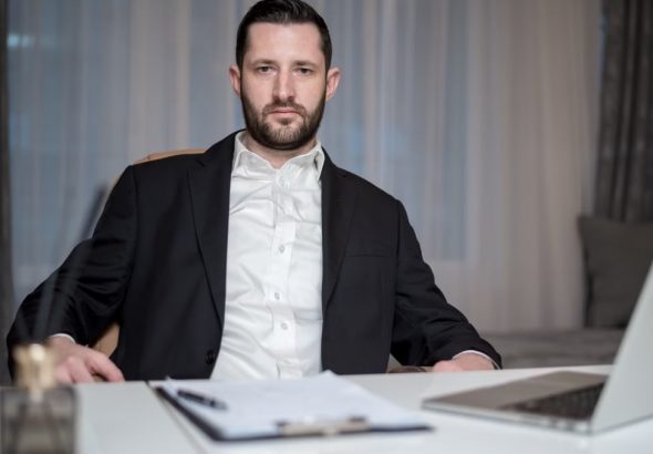 A serious bearded man in a black suit sits behind a desk with a laptop and documents.