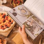 A person reads a cookbook while preparing a fruit tart on a wooden table.