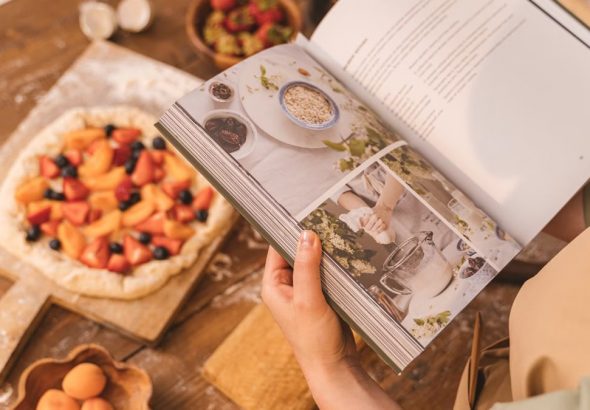 A person reads a cookbook while preparing a fruit tart on a wooden table.